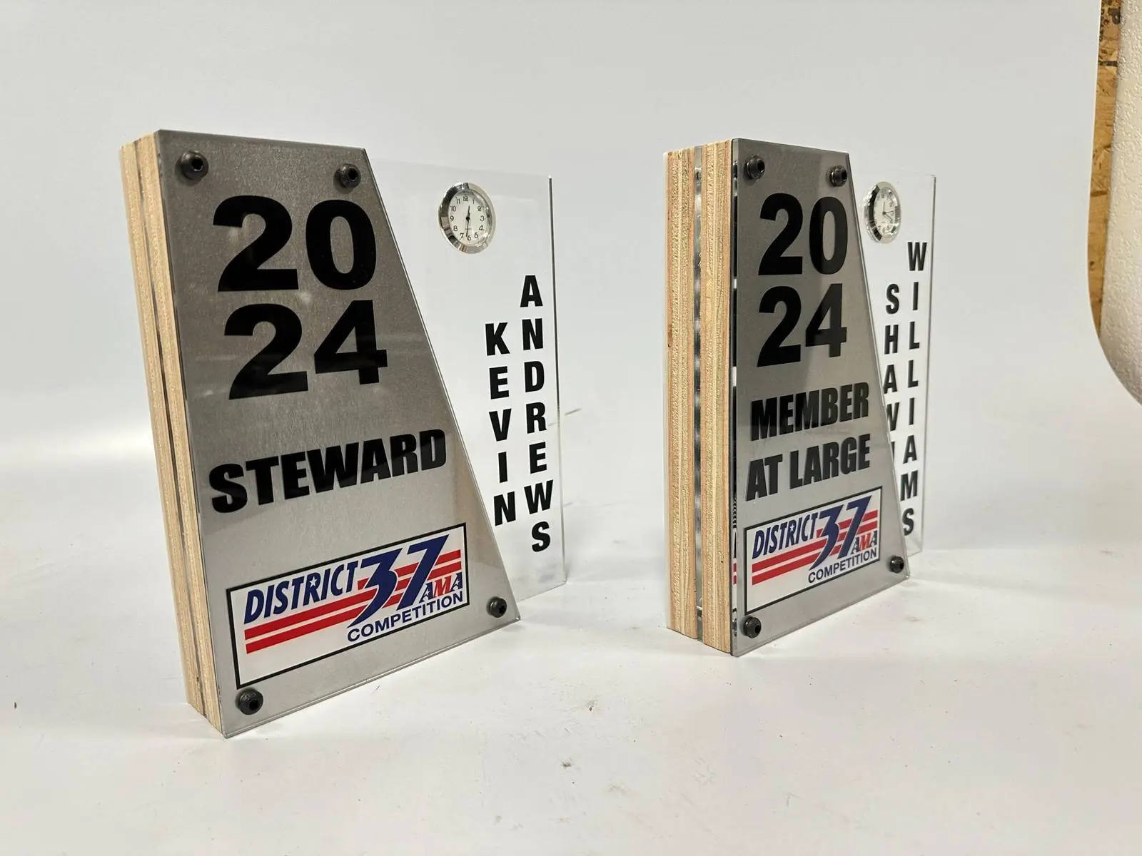 Two competition committee awards, each a brushed-finish plate on a wood and clear acrylic body with the recipient’s name set vertically