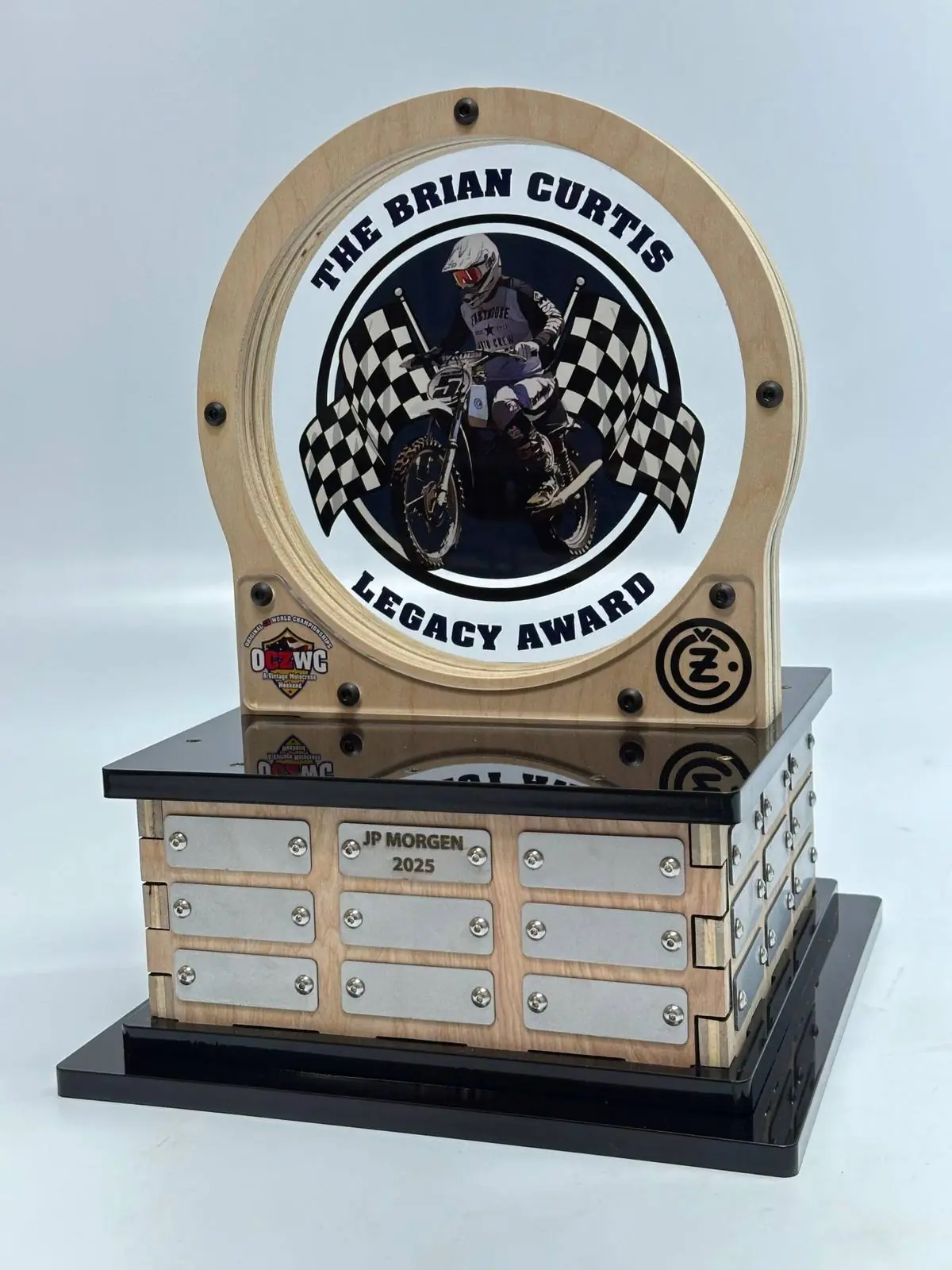 A perpetual award with a printed rider graphic in a wood ring above a base carrying nine engraved name plates, the top one filled in and the rest blank