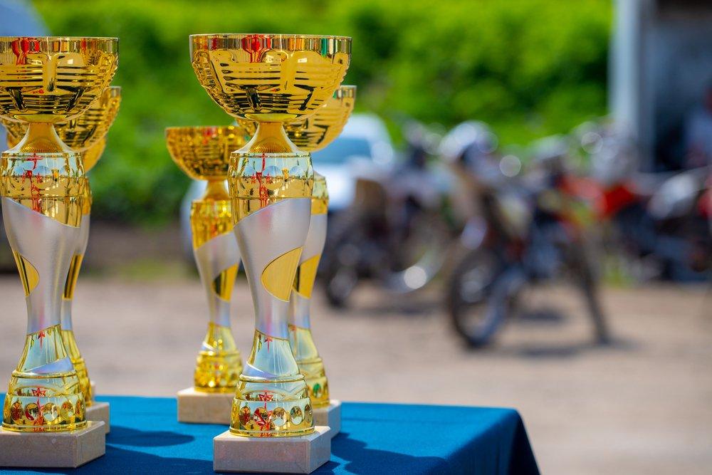Gold and silver racing trophies on a blue-draped table outdoors with motorcycles parked behind