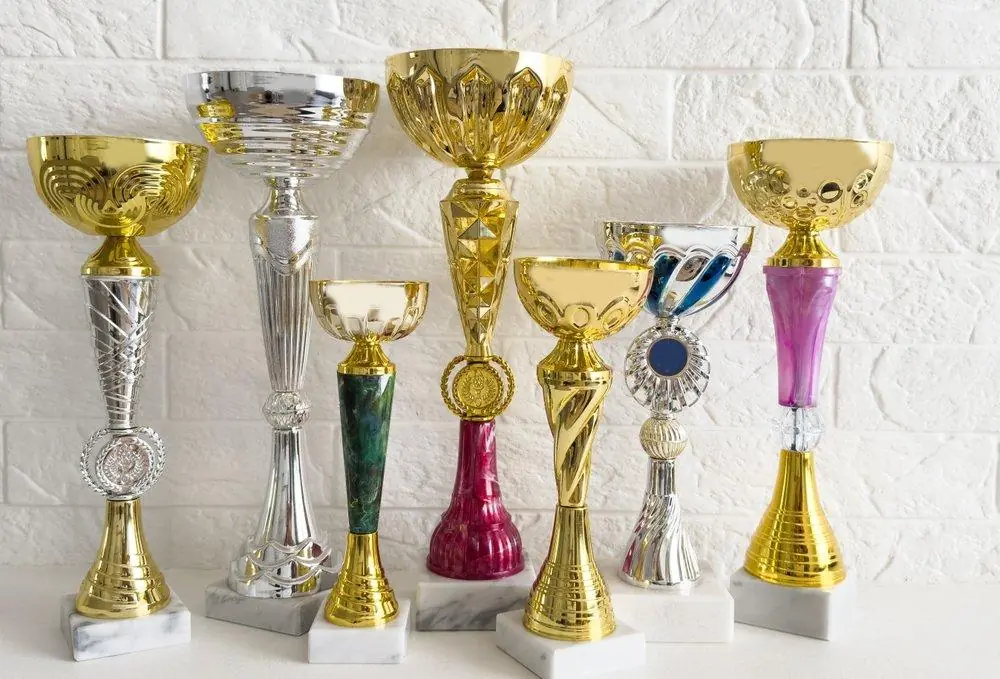 Seven gold and silver cup trophies of different heights on marble bases against a white brick wall
