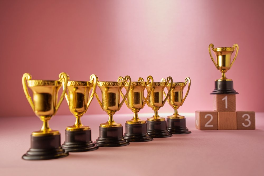 Six small gold cup trophies in a row beside a wooden first, second and third place podium