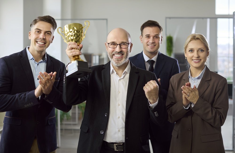 Colleagues applauding as a man holds up a gold cup trophy in an office